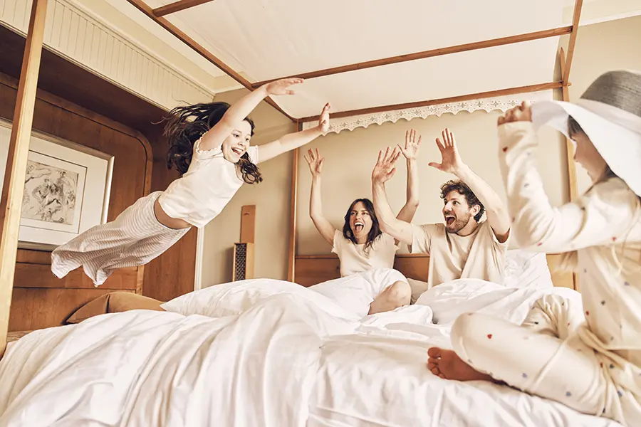 Ein Kind springt mit ausgestreckten Armen auf einem Bett, während eine Familie in Pyjamas begeistert zuschaut. Das Zimmer hat Holzmöbel und eine gemütliche Atmosphäre und befindet sich in einem Hotel in Vaterstetten.